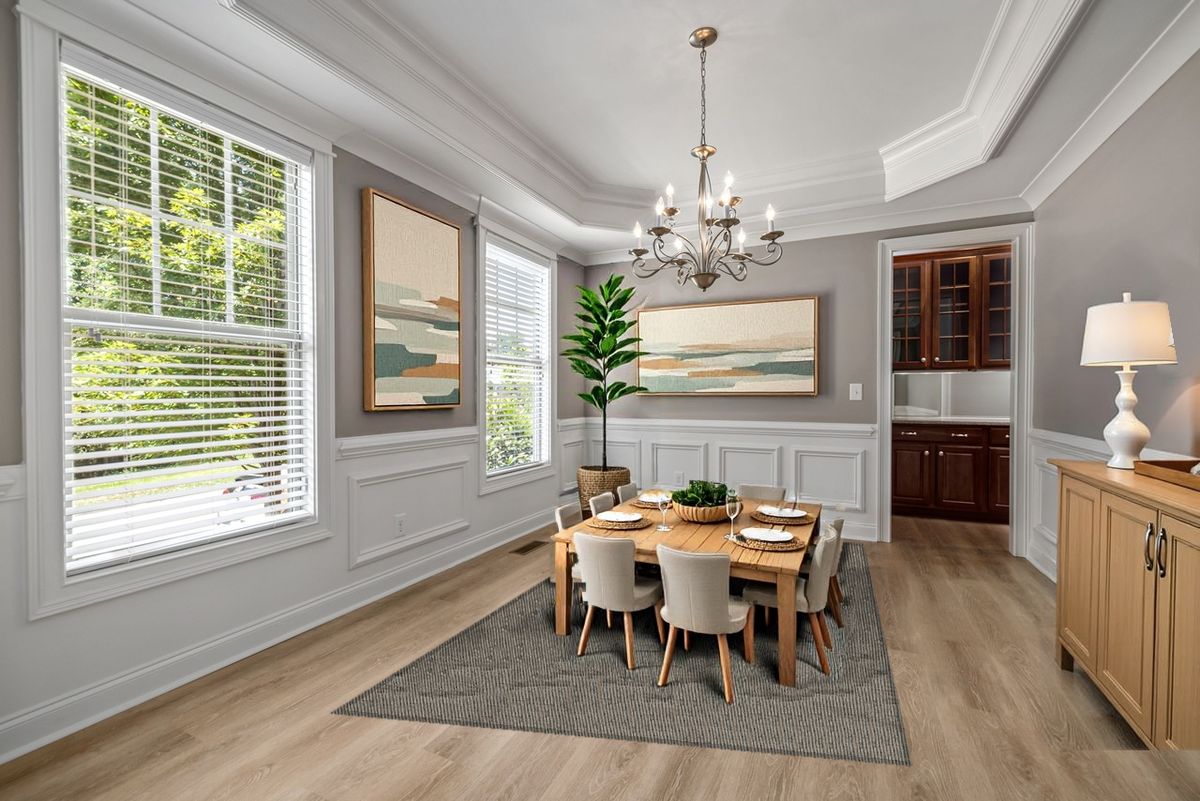Virtually staged: the same dining room with a digitally added oak table and eight upholstered chairs, place settings, woven rug, two framed paintings, a fiddle-leaf fig and an oak buffet with a lamp; walls, windows, chandelier and floor unchanged
